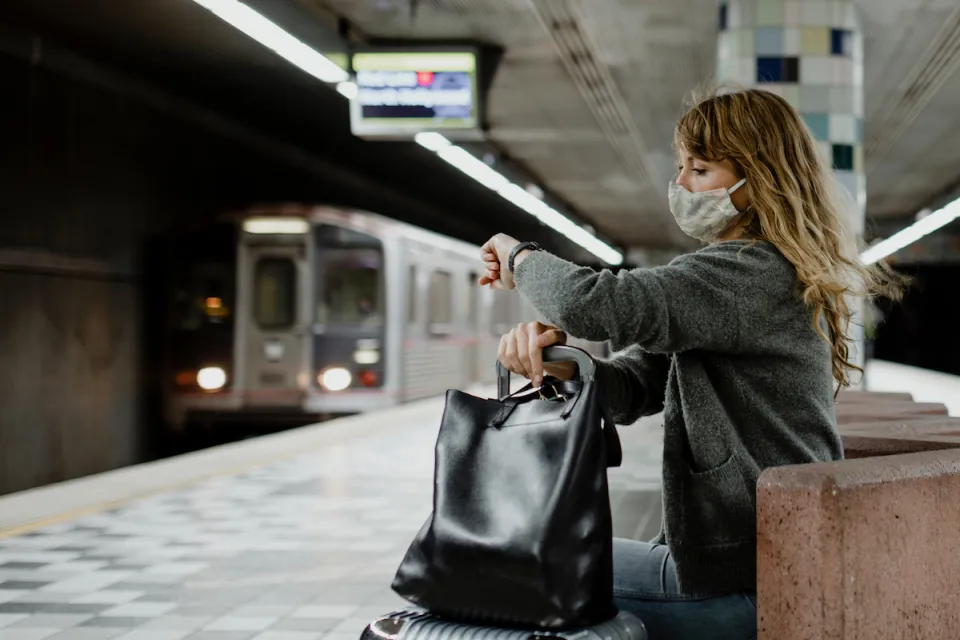 Woman with long hair wearing Covid mask sitting on underground train station bench looking at her watch as subway train approaches in background at Hollywood/Western station in Los Angeles, California.
